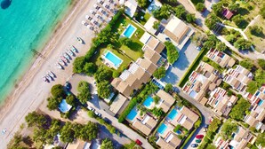 Oversigtsbillede af en kystresort med overnatningsmuligheder ved stranden, en swimmingpool og krystalklart hav.