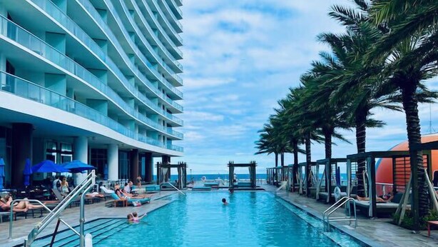 A swimming pool with lounge chairs and umbrellas, surrounded by palm trees and a hotel building.