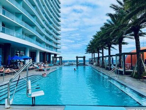 En swimmingpool med solstolar och parasoller, omgiven av palmer och en hotellbyggnad.