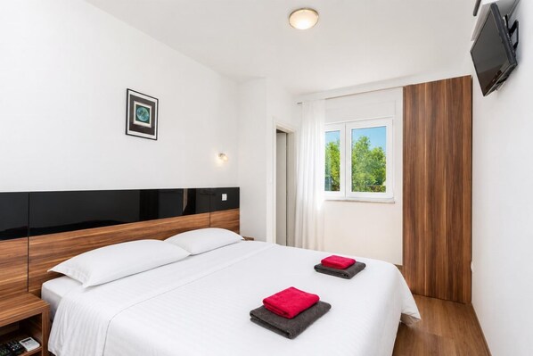 A modern hotel room with a large bed, a bedside table, a flat-screen TV mounted on the wall, and a window with a view of greenery.