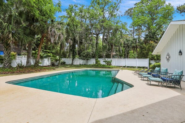 A rectangular swimming pool with a white deck, surrounded by lush greenery and trees.