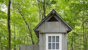 Una pequeña construcción de madera de un solo piso con una puerta y ventanas blancas, rodeada por un camino de losetas de piedra y una valla de madera, ubicada en un área boscosa.