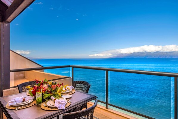 A dining table set with plates, cutlery, and a floral centerpiece on a balcony with a view of the ocean and mountains.