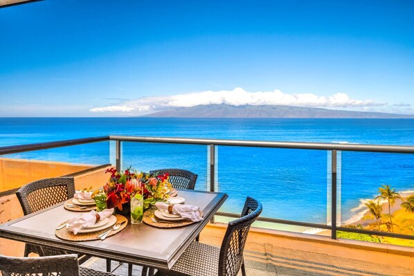 A table set with plates, cutlery, and a vase of flowers on a balcony with a view of the ocean and a mountain range.