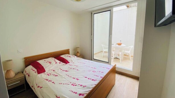 A bedroom with a bed, a nightstand, a lamp, a sliding glass door leading to a balcony with a table and chairs. - Appât T2 en Rdc à 50 Mètres de la Plage (Saint Palais sur Mer)