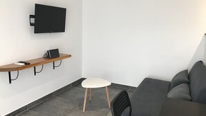 Een moderne, compacte leefruimte met een flatscreen televisie aan de muur, een houten plank met een laptop en afstandsbedieningen, een witte stoel, een grijze bank en een betegelde vloer.