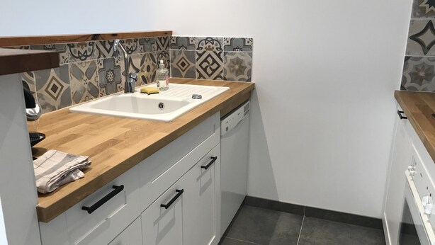 Een compacte keuken met een houten werkblad, witte keukenkastjes, een spoelbak en een betegelde spatwand met een patroon.