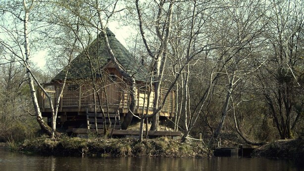 A rustic cabin by a river with a wooden deck and a thatched roof. - Cabane : île aux Canets, Thoureil, Maine & Loire (Gennes-Val-de-Loire)