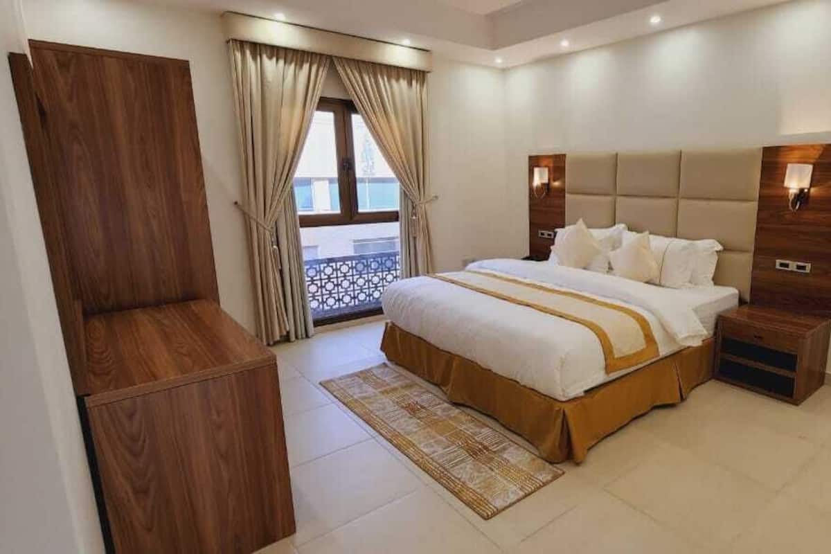 A modern hotel room with a large bed, wooden headboard, bedside tables, and a view of the outdoors through a window.