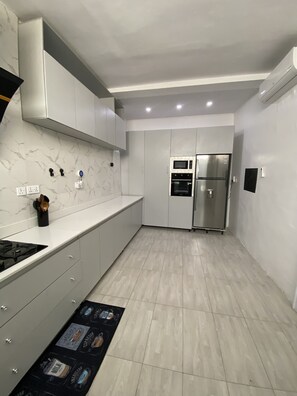 Una cocina moderna con gabinetes grises, microondas integrado, horno y refrigerador. Hay una estufa a gas y un tapete negro con imágenes en el suelo.