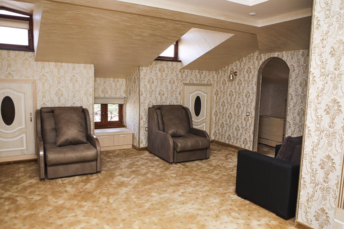 Ein geräumiges Zimmer mit zwei Ledersesseln, einem schwarzen Sofa, ornamentalem Wandpapier und einem Oberlicht.