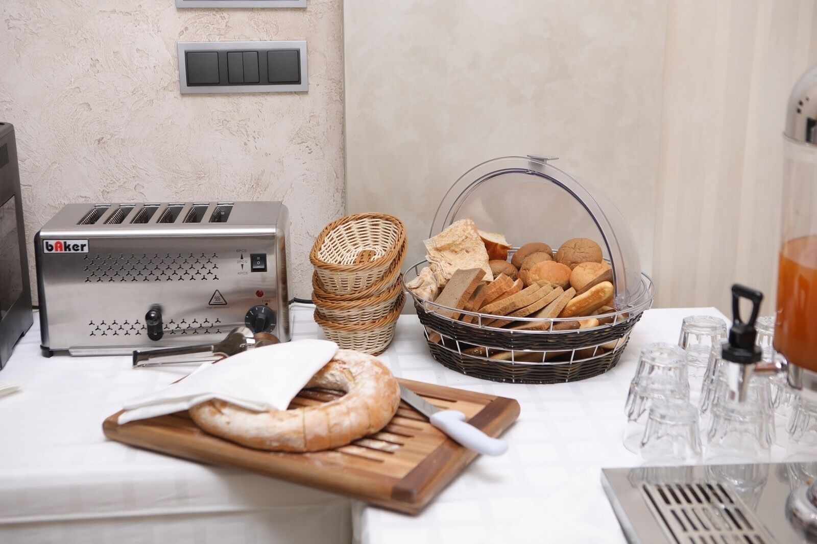 A kitchen area with a bread-making machine, a basket of bread, a cutting board with a loaf of bread, and a tea dispenser.
