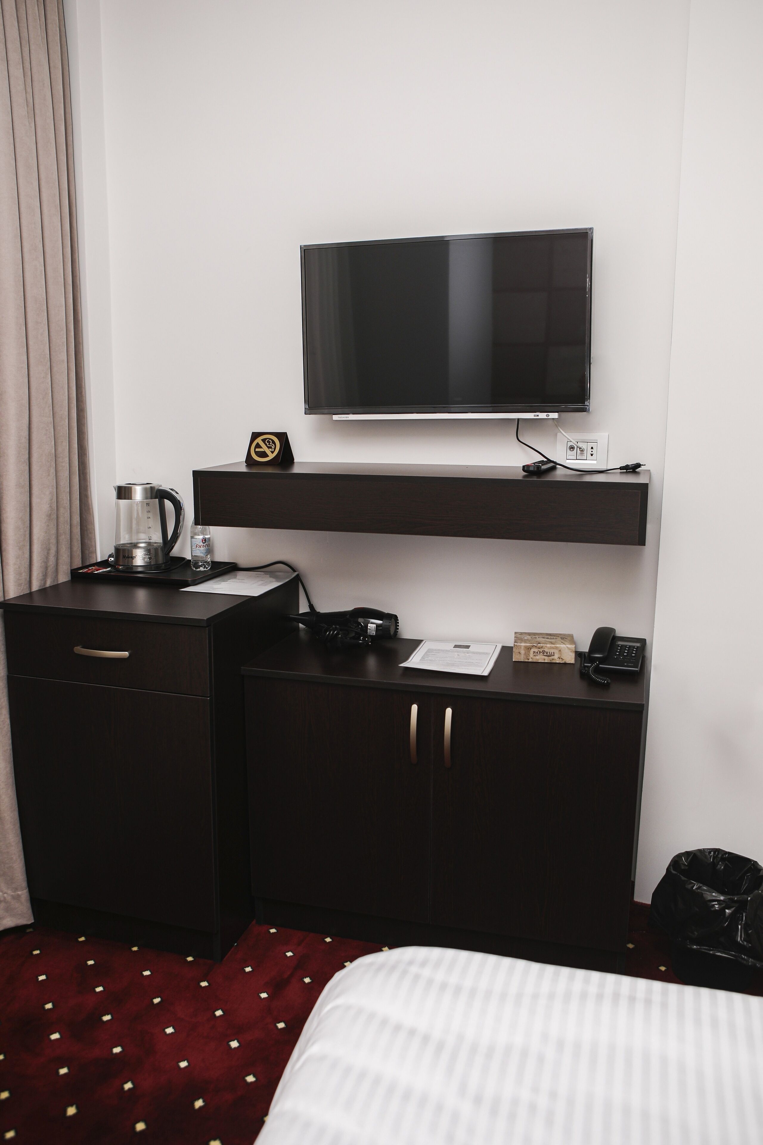 Ein Hotelzimmer mit Bett, Schreibtisch mit Kaffeemaschine, Telefon, Wandfernseher und kleiner Uhr.