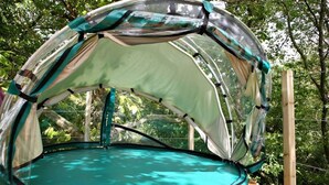 A dome-shaped tent with a transparent floor, set up outdoors with trees in the background.