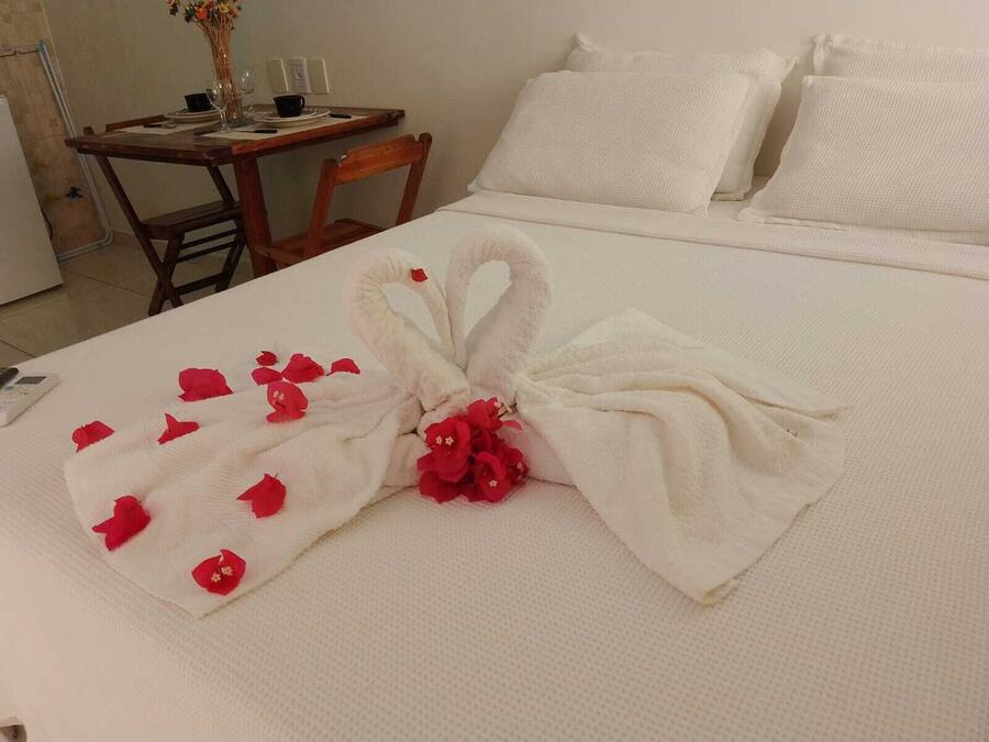 A neatly made bed with a towel swan centerpiece, white bedding, and a small table with a vase of flowers.