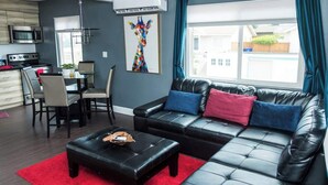 Een moderne woonkamer met een zwarte leren bank, een eettafel met stoelen, een rode vloerkleed en een groot raam met gordijnen.