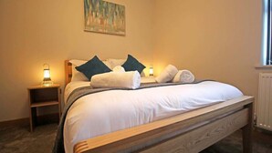 Cama bem arrumada com lençóis brancos e almofadas escuras, luminária de cabeceira e criado-mudo de madeira.