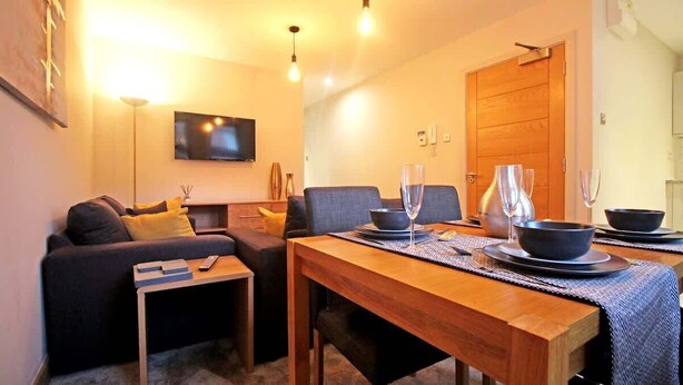 Uma sala de estar aconchegante com um sofá, uma mesa de jantar de madeira posta com tigelas e copos, e uma TV fixada na parede.