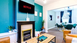 A living room with a fireplace, a TV mounted on the wall, and a dining area with green chairs.