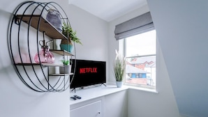 Moderni olohuone, jossa on valkoinen seinä, johon on asennettu televisio, metallinen hylly, jossa on kasveja, ja ikkuna, josta avautuu näkymä rakennuksiin.
