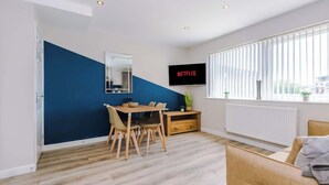 A modern living room with a dining area, a TV mounted on the wall, and a large window with blinds. - Hidden Gem within Central Chester - Great Location (Chester)