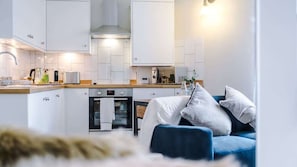 Une cuisine moderne avec des armoires blanches, un four encastré et un four à micro-ondes. Au premier plan, on aperçoit un canapé bleu avec des coussins blancs.