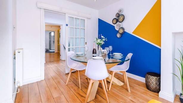 Un coin repas avec une table en verre, des chaises blanches et un mur bleu et jaune.