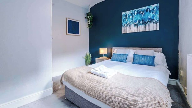 A modern bedroom with a large bed, a nightstand, a wall-mounted painting, and a potted plant.