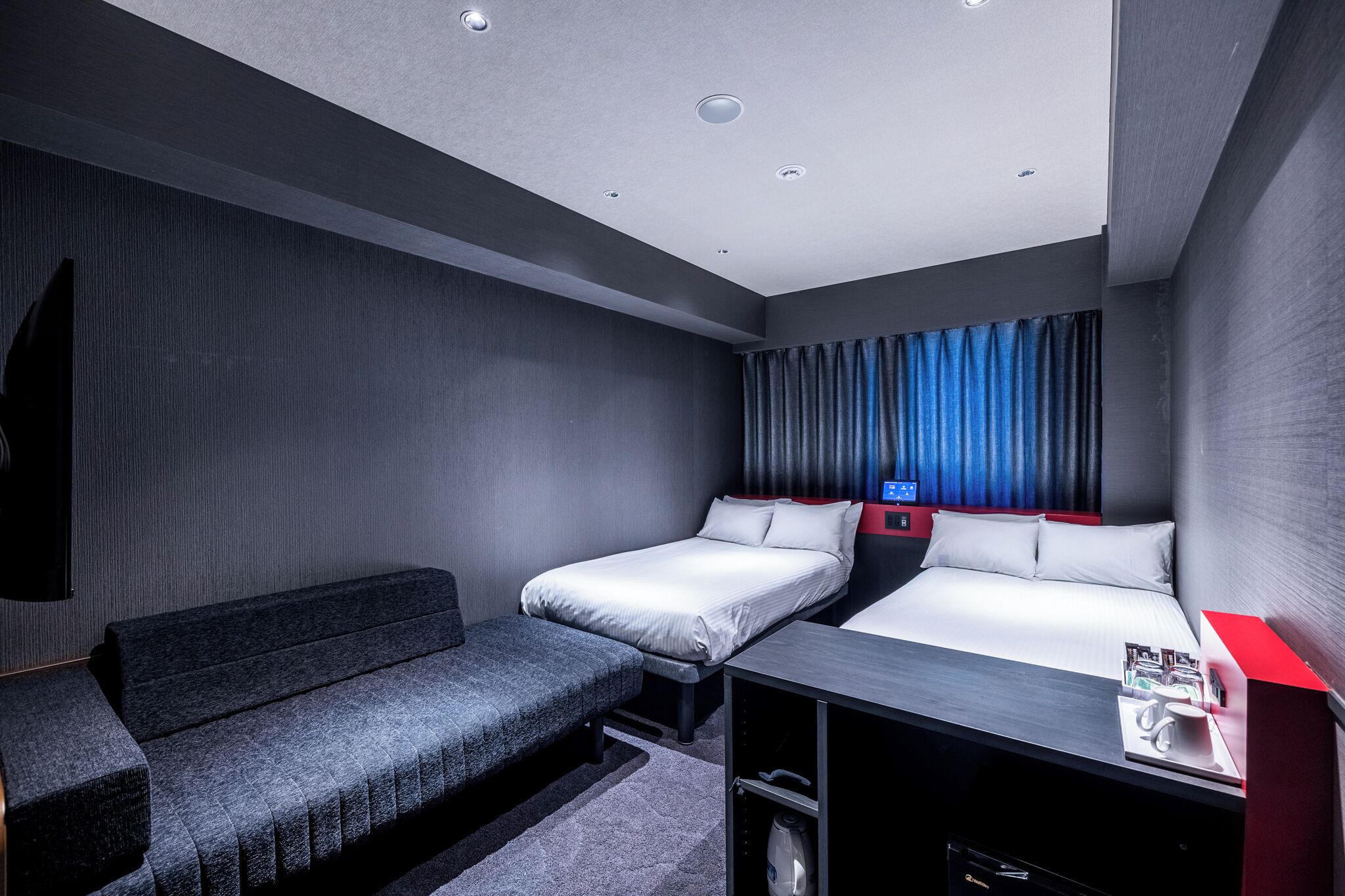 A modern hotel room with a sofa, two single beds, a desk with a red accent, and a TV mounted on the wall.