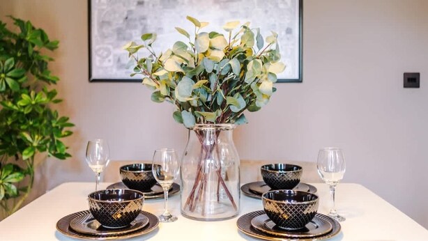 Une table à manger dressée avec des bols noirs et dorés à motifs, des verres à vin en verre transparent et un vase transparent contenant des feuilles vertes.