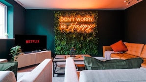 Un salon moderne avec un néon affichant le message « NE VOUS INQUIÉTEZ PAS, SOYEZ HEUREUX », un téléviseur diffusant Netflix et un mur vert orné de plantes.