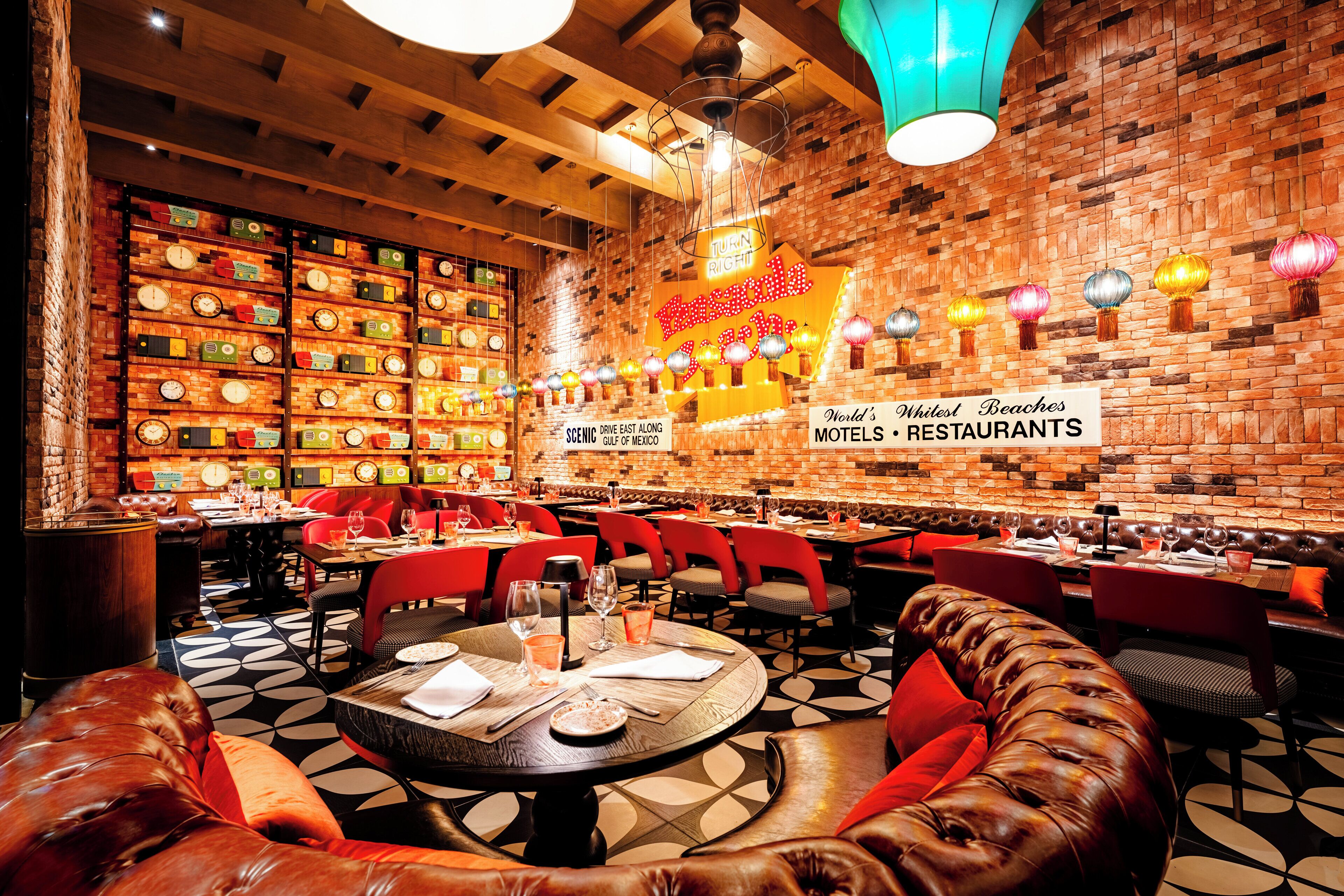 L’intérieur d’un restaurant avec des murs en briques, de nombreuses tables, des chaises rouges et un panneau lumineux au néon.