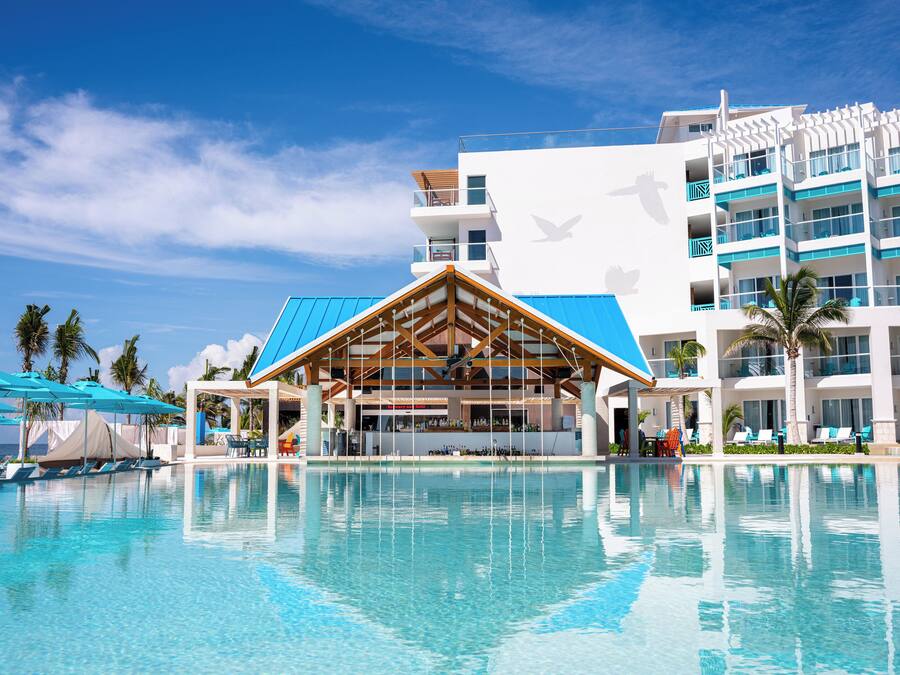Un resort con una piscina grande, una zona de bar techada y un edificio de varios pisos con balcones.
