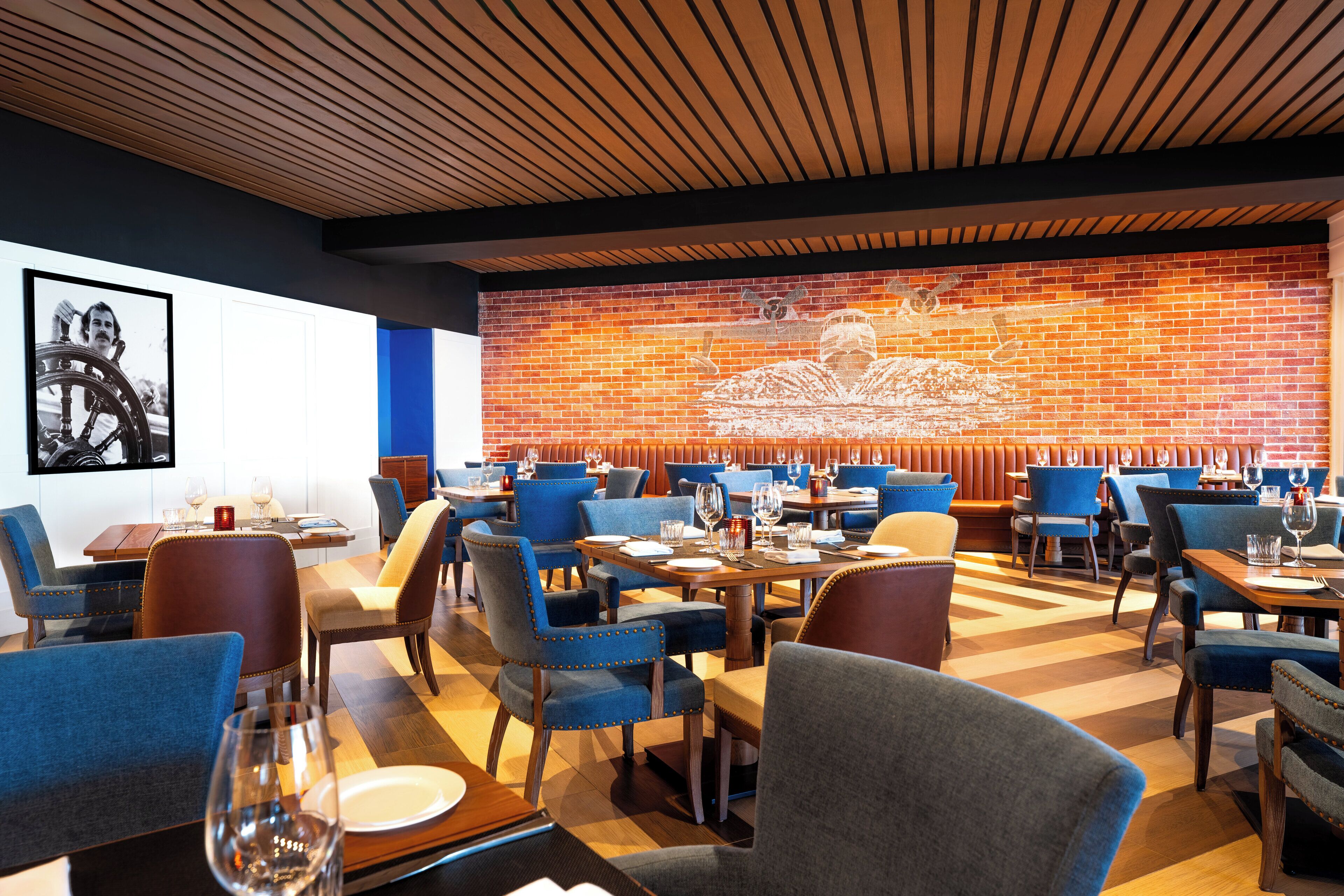 Un restaurant avec des chaises à revêtement bleu, des tables en bois et un mur de briques orné d’une fresque.