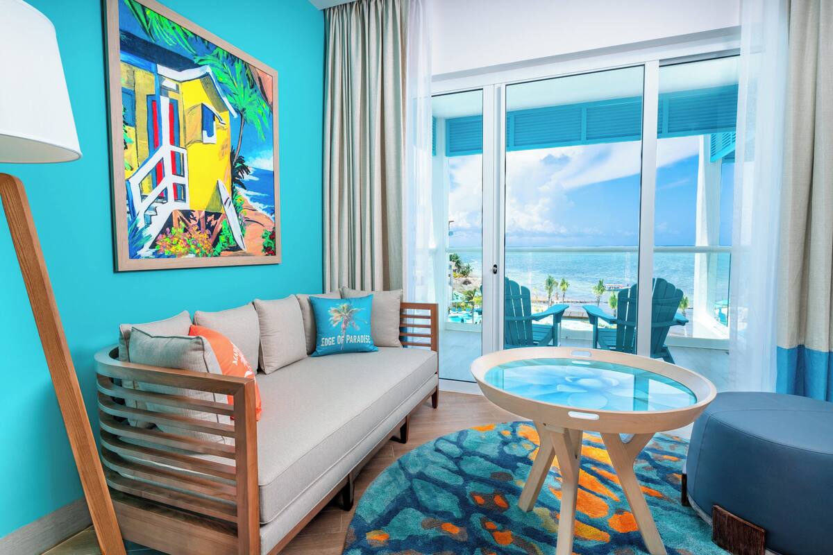 A room with a sofa, a glass-top table, a view of the ocean, and a painting on the wall.