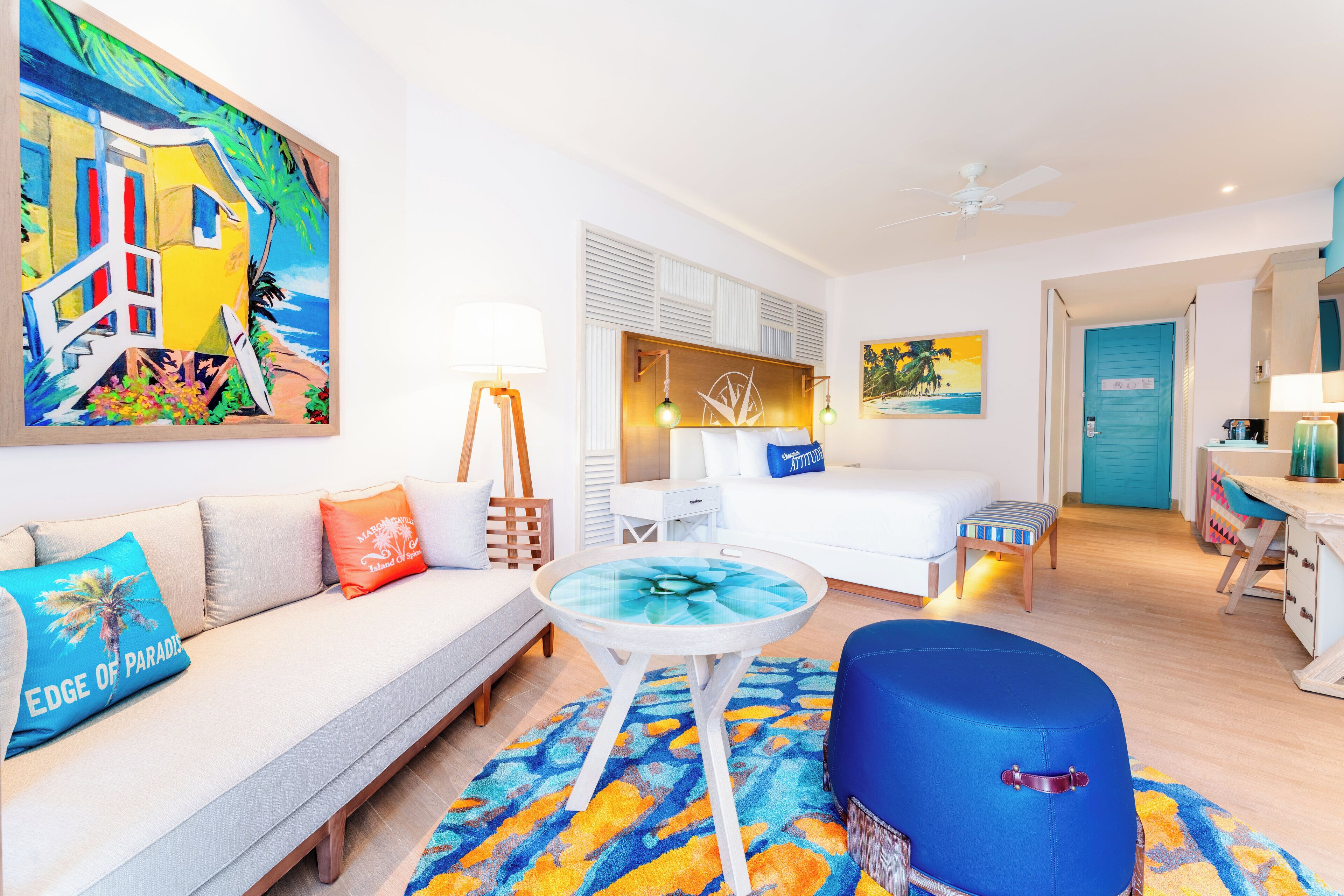 Une chambre d’hôtel avec un lit, un canapé, une petite table, un pouf bleu et un tableau représentant une scène de plage.