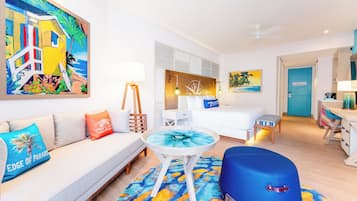 Une chambre d’hôtel avec un lit, un canapé, une petite table, un pouf bleu et un tableau représentant une scène de plage.