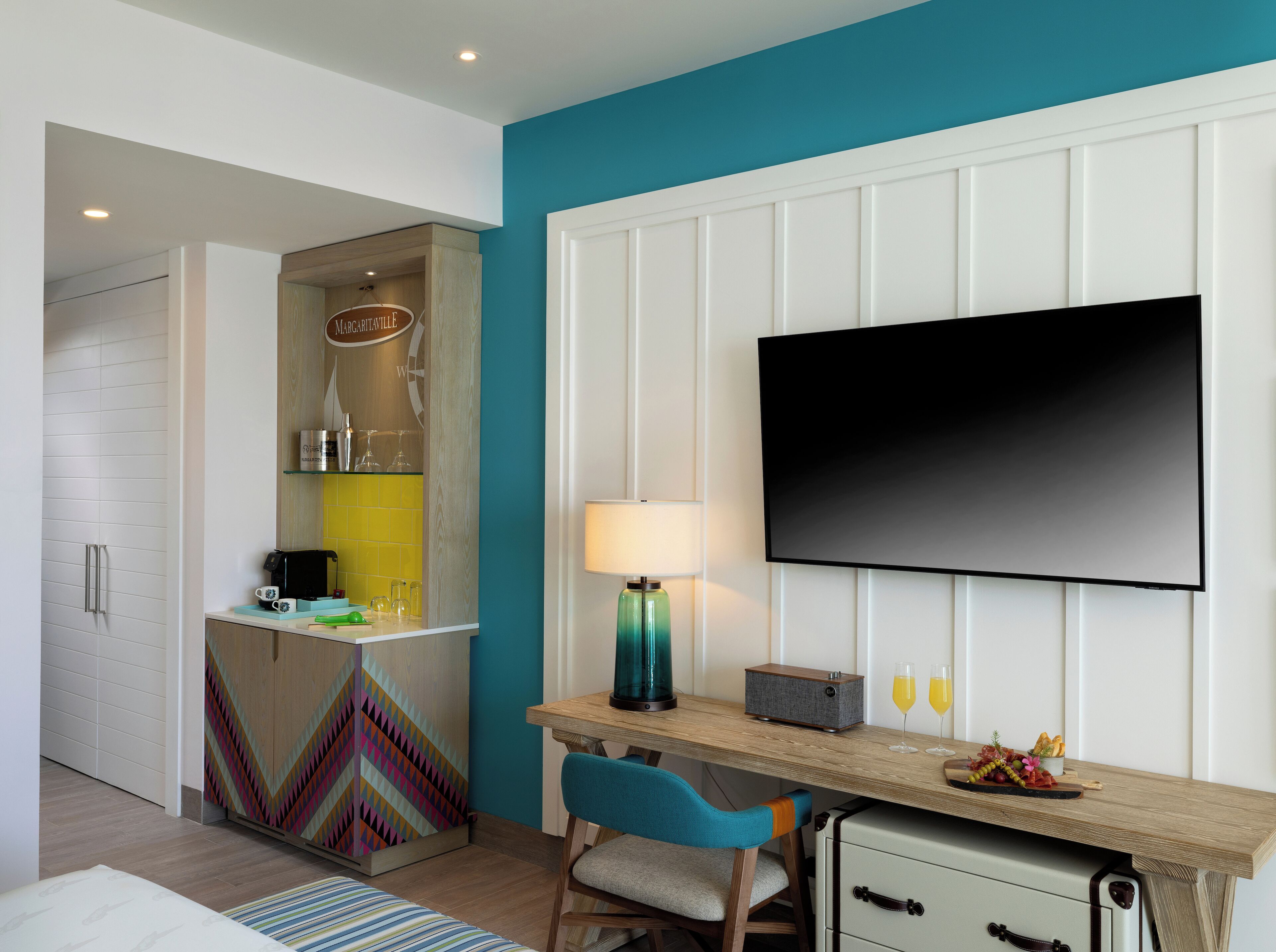 Een moderne hotelkamer met een kitchenette, een flatscreen televisie bevestigd aan een witte muur, een bureau met een lamp en een stoel.