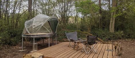 A camping setup with a tent, two chairs, and a wooden deck in a forest.