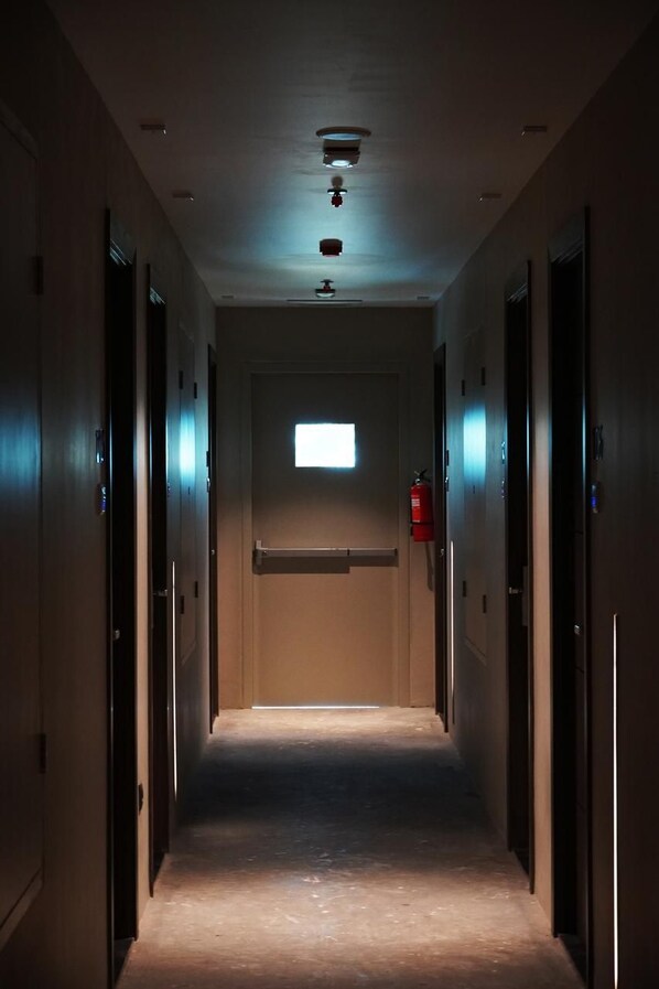 A narrow hallway with doors on both sides, a fire extinguisher on the right, and a small window at the end.