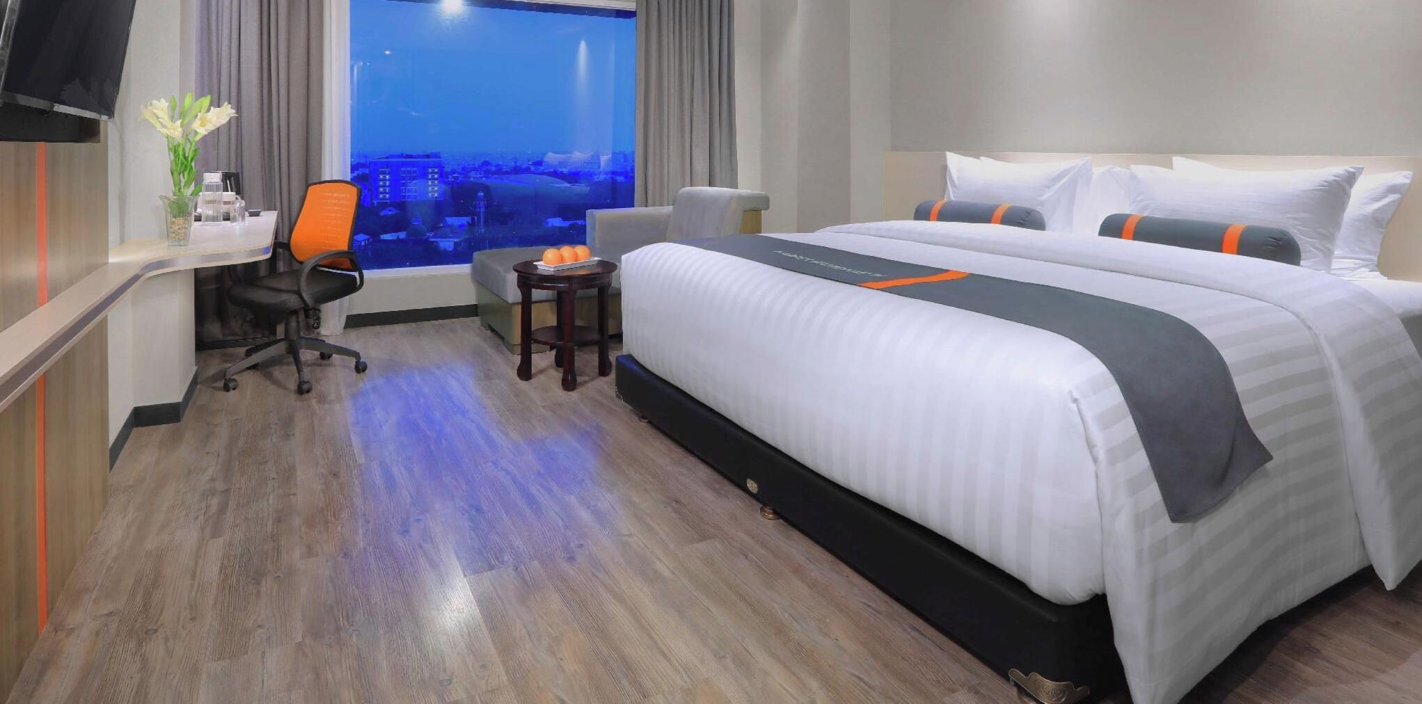 A hotel room with a large bed, a desk with a chair, a small table, and a view of a cityscape through a window.