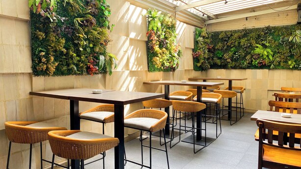 En restaurang med högt i tak, naturligt ljus och vertikala trädgårdar på väggarna. Bord och stolar av rotting är placerade ut för att skapa en trevlig matplats.