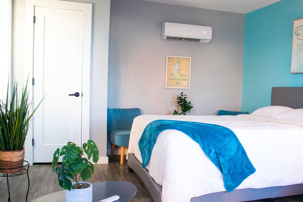 A hotel room with a bed, a bedside table with a lamp, a wall-mounted air conditioner, a framed inspirational quote, and a potted plant.