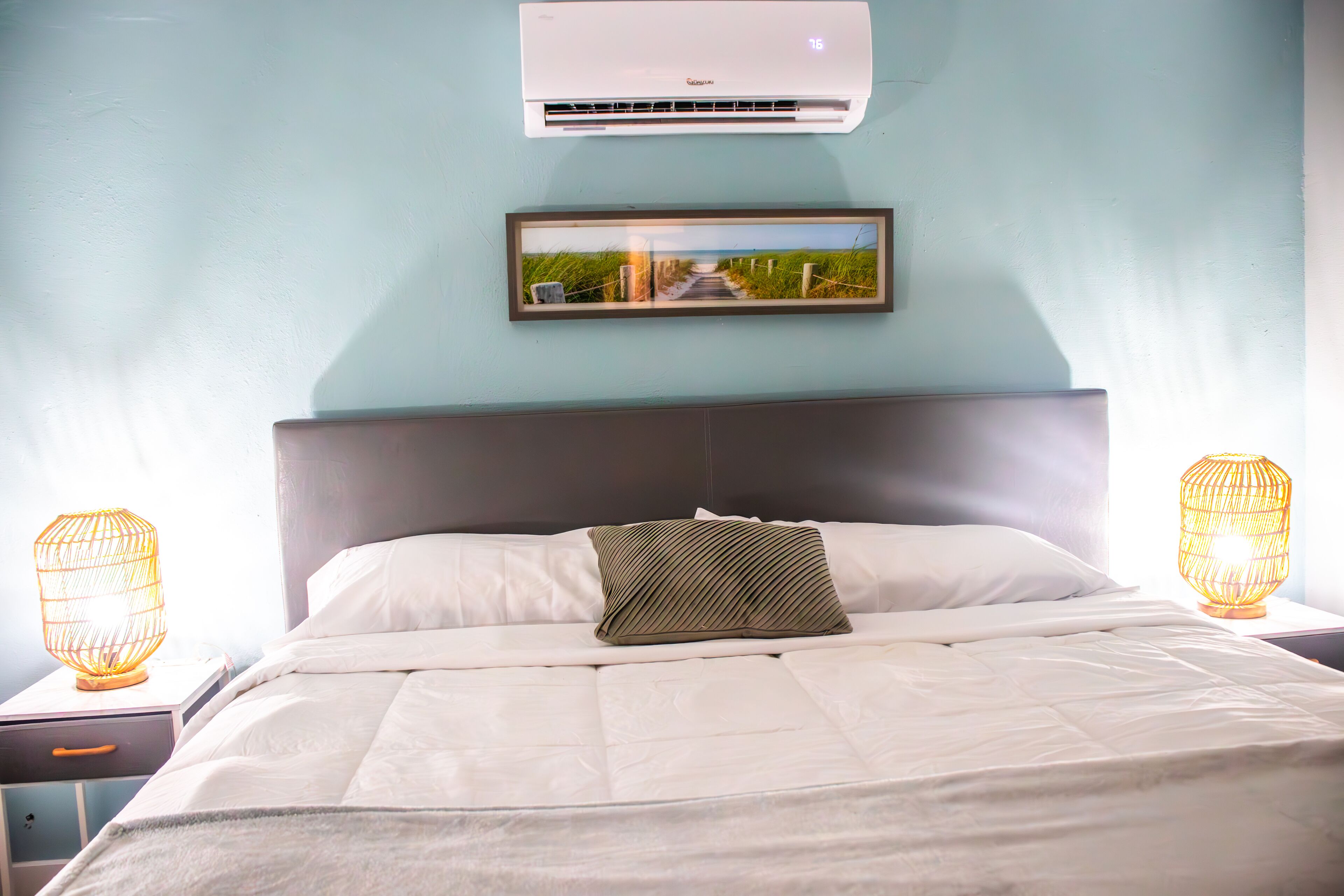 A neatly made bed with a white comforter and a single patterned pillow, a framed picture above the bed, and an air conditioner mounted on the wall.