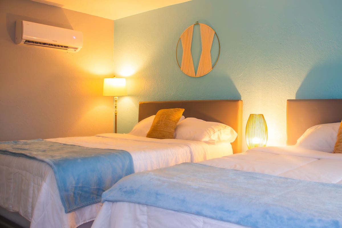 A hotel room with two beds, a wall-mounted air conditioner, and a bedside lamp.