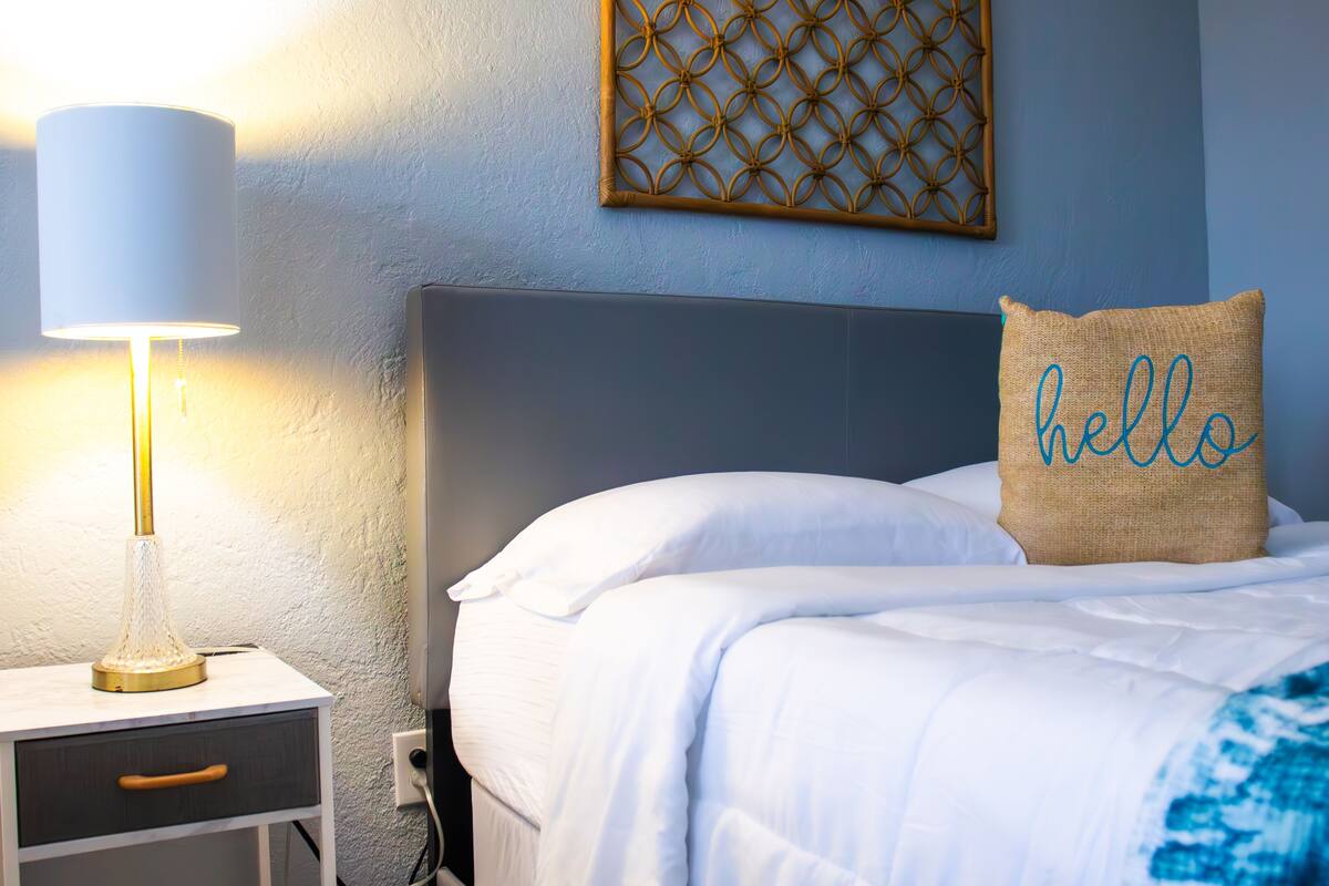A neatly made bed with white linens, a "hello" pillow, a bedside lamp, and a small wooden nightstand with a drawer.
