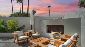 An outdoor patio with wooden furniture, a fire pit, and a view of palm trees against a sunset sky.