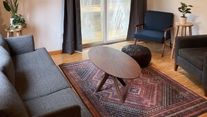 Ein Wohnzimmer mit Holzbalkendecke, einem großen Fenster mit Vorhängen, einem blauen Sessel, einem grauen Sofa, einem kleinen runden Tisch und einem gemusterten Teppich.