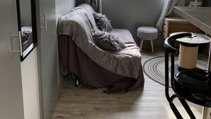 Een kleine slaapkamer met een bed, een raam, een klein krukje en een krabpaal.
