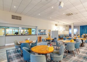 A modern hotel lobby with round tables and blue chairs, a bar area, and a seating area with a blue curtain backdrop.